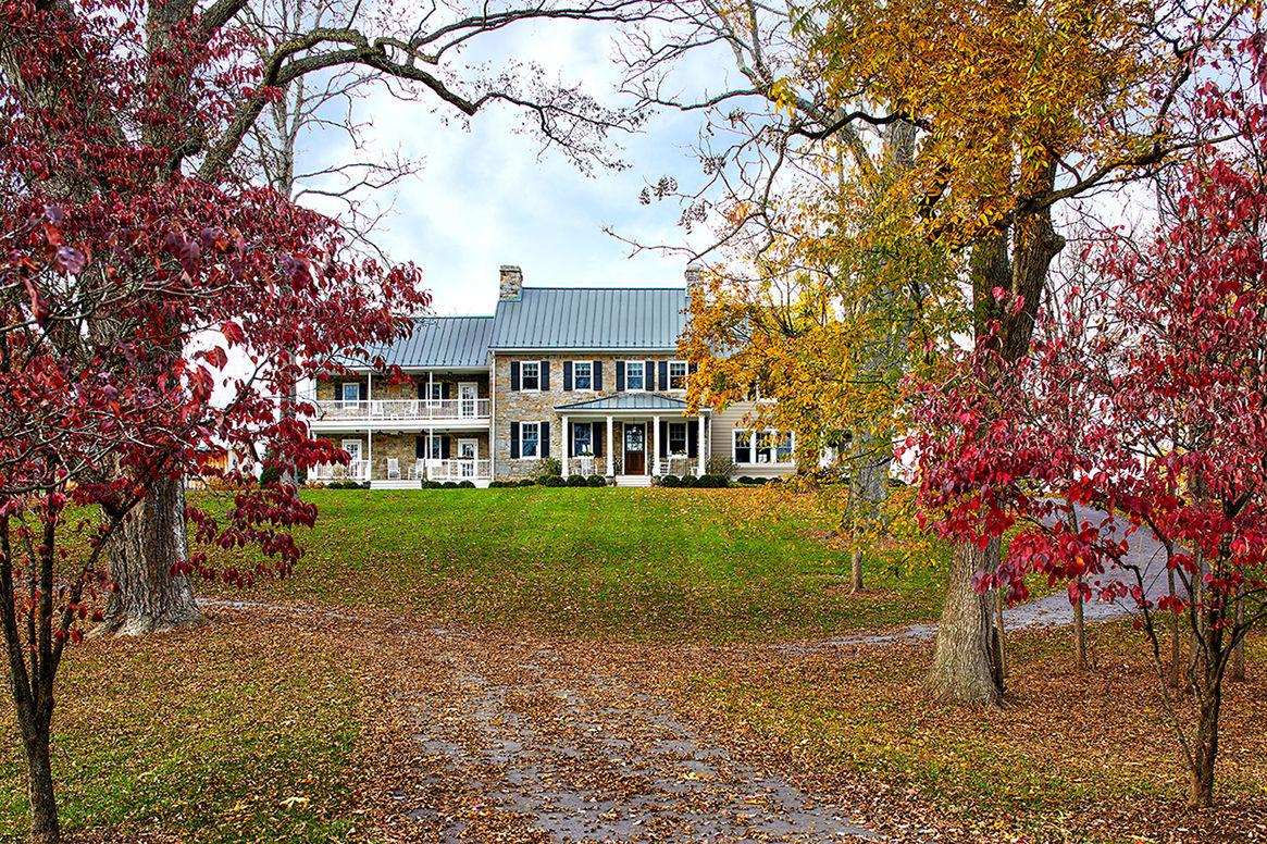 Traditional stone farmhouse with metal roof and symmetrical façade, featuring double porches with white columns and rocking chairs, set on a grassy hill framed by vibrant autumn trees.